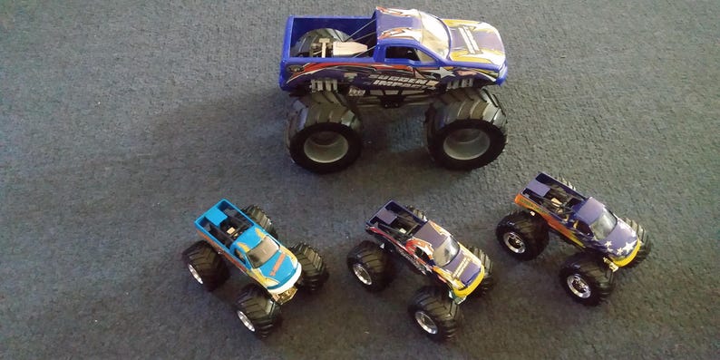 K&ouml;nnte beinhalten: Eine Sammlung von Spielzeug-Monstertrucks in verschiedenen Farben, darunter Blau, Gelb und Lila. Die Trucks haben gro&szlig;e, schwarze Reifen und detaillierte Designs. Ein Truck ist gr&ouml;&szlig;er als die anderen.