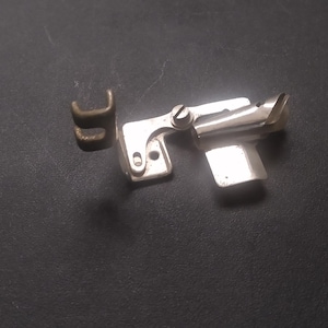 May include: A silver-colored sewing machine attachment with a curved edge and a small, separate brass-colored piece. The attachment has a screw and a flat, rectangular piece. The items are on a dark surface.