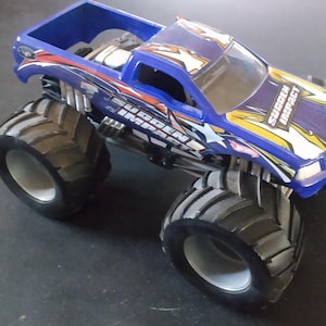 K&ouml;nnte beinhalten: Ein blaues Spielzeug-Monstertruck mit gro&szlig;en schwarzen Reifen. Der Truck hat eine blaue Kabine und Ladefl&auml;che mit Grafiken, die "Sudden Impact" lauten. Der Truck steht auf einer dunklen Oberfl&auml;che.