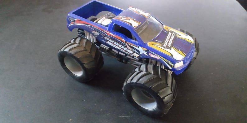 K&ouml;nnte beinhalten: Ein blaues Spielzeug-Monstertruck mit gro&szlig;en schwarzen Reifen. Der Truck hat eine blaue Kabine und Ladefl&auml;che mit Grafiken, die "Sudden Impact" lauten. Der Truck steht auf einer dunklen Oberfl&auml;che.