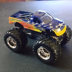 K&ouml;nnte beinhalten: Ein Spielzeug-Monstertruck mit gro&szlig;en schwarzen Reifen und einer blau, orange und wei&szlig;en Karosserie. Der Truck hat den Schriftzug "SUDDEN IMPACT" an der Seite und wei&szlig;e Sterne. Die Vorderseite des Trucks ist gelb.