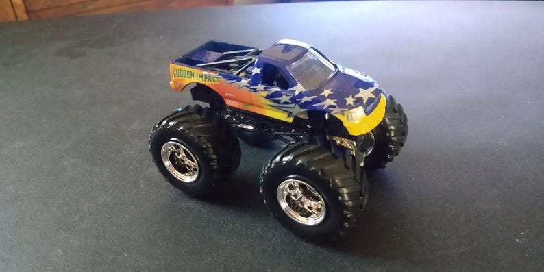 K&ouml;nnte beinhalten: Ein Spielzeug-Monstertruck mit gro&szlig;en schwarzen Reifen und einer blau, orange und wei&szlig;en Karosserie. Der Truck hat den Schriftzug "SUDDEN IMPACT" an der Seite und wei&szlig;e Sterne. Die Vorderseite des Trucks ist gelb.