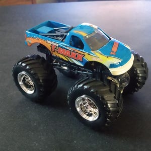 K&ouml;nnte beinhalten: Ein blau-oranger Spielzeug-Monstertruck mit dem Text "T-MAXX" an der Seite. Der Truck hat gro&szlig;e schwarze Reifen mit silbernen Felgen. Der Truck ist eine Miniatur-Nachbildung eines Monstertrucks.