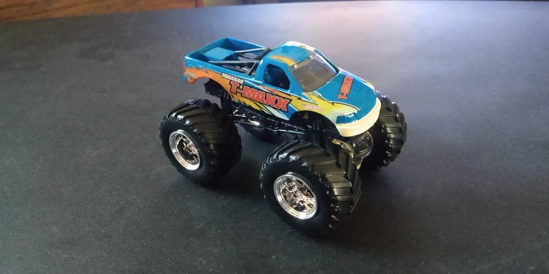 K&ouml;nnte beinhalten: Ein blau-oranger Spielzeug-Monstertruck mit dem Text "T-MAXX" an der Seite. Der Truck hat gro&szlig;e schwarze Reifen mit silbernen Felgen. Der Truck ist eine Miniatur-Nachbildung eines Monstertrucks.