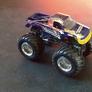 K&ouml;nnte beinhalten: Ein blau-gelbes Monstertruck-Spielzeug mit gro&szlig;en schwarzen Reifen und silbernen Felgen. Der Truck hat ein detailliertes Design mit Sterngrafiken und dem Text "Sudden Impact".