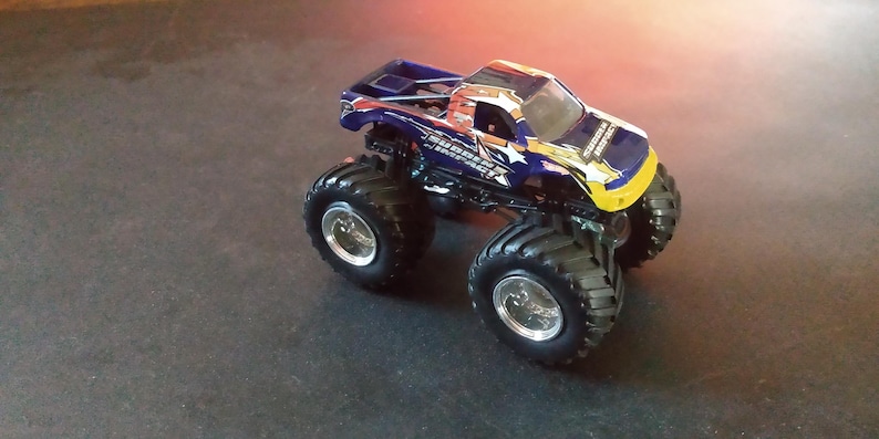 K&ouml;nnte beinhalten: Ein blau-gelbes Monstertruck-Spielzeug mit gro&szlig;en schwarzen Reifen und silbernen Felgen. Der Truck hat ein detailliertes Design mit Sterngrafiken und dem Text "Sudden Impact".