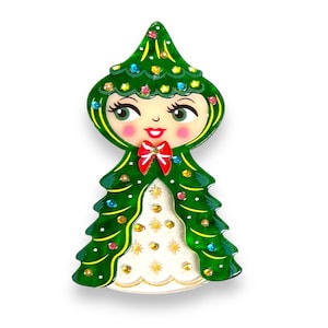 May include: A whimsical Christmas tree-shaped brooch. The brooch features a smiling face with rosy cheeks, green eyes, and a red bow. The green tree design is adorned with colorful gems and a white dress with gold accents.