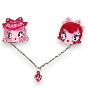 Peut inclure: Broche fantaisiste avec deux visages stylisés, un rose et un rouge, avec des nœuds et de grands yeux, reliés par une chaîne argentée. Un charm en forme d'ornement rouge est suspendu à la chaîne. Sur fond blanc.