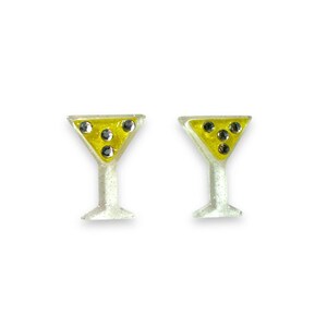 Op de afbeelding: Een paar cocktail-thema oorbellen. Elke oorbel heeft de vorm van een martiniglas met een gele vloeistof en zwarte edelsteenaccenten. De stelen en bases zijn wit met glitter.