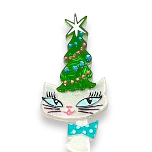 Peut inclure: Broche fantaisie sur le thème de Noël. Un chat blanc porte un sapin vert orné de boules bleues et roses. Un nœud papillon turquoise à pois blancs complète le design. La broche est faite d'un matériau brillant.