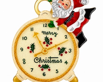 May include: A gold-toned metal brooch shaped like an alarm clock with a cream-coloured face, green numbers, and holly leaf accents. The text reads merry Christmas. A Santa Claus figure in a red and white suit is attached to the top right side.