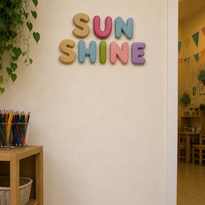 Könnte beinhalten: An einer cremefarbenen Wand sind bunte Holzbuchstaben mit der Aufschrift "SUN SHINE" angebracht. Die Buchstaben sind in Gelb-, Rosa-, Blau-, Grün- und Lilatönen gehalten. Im Hintergrund ein Holzregal mit Kunstbedarf und eine Hängepflanze.
