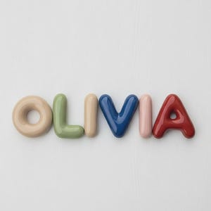 May include: Colorful, 3D letters spelling "OLIVIA" in various shades. The letters are in a playful, rounded font, with colors including beige, green, tan, blue, pink, and red. The letters are arranged on a white background.