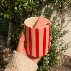 May include: A ceramic mug with a lid, featuring a pink and red striped design. The mug is cylindrical with a rounded top and a lid with a small hole. The mug is held in a person's hand, with a green leafy background.