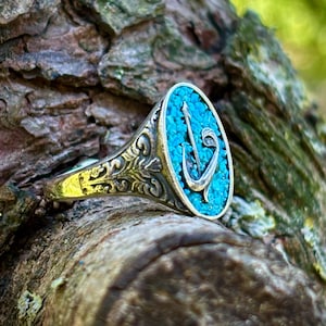 May include: A silver ring with an oval turquoise stone inlay. The stone features a silver anchor design. The ring has an ornate, engraved band. The ring is set against a textured, brown tree trunk.