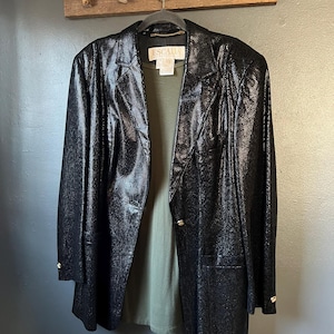 May include: A black blazer with a textured snakeskin-like pattern, featuring a single button closure, notched lapels, and long sleeves with button cuffs. An olive green shirt is layered underneath, and an Escada label is visible at the neck.