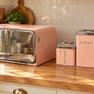 May include: A pastel pink bread box with a reflective silver door and three matching canisters labeled COFFEE, SUGAR, and TEA. Each item features a diagonal ribbed texture and a metallic silver lid with a small circular knob on top.