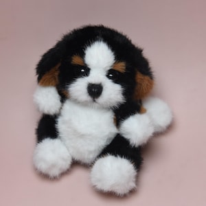 May include: A plush toy puppy with a black, white, and brown coat. The stuffed animal has a white face with black eyes and a black nose. The ears are brown, and the paws are white. The toy is on a pink background.