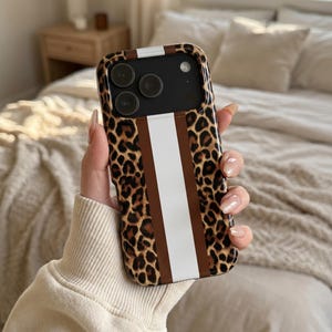 Könnte beinhalten: Eine Smartphone-Hülle mit braun-schwarzem Leopardenmuster und einem vertikalen weißen Streifen in der Mitte. Die Hülle hat einen Ausschnitt für die Kameralinsen und eine glänzende Oberfläche, die die Rückseite des Mobilgeräts bedeckt.