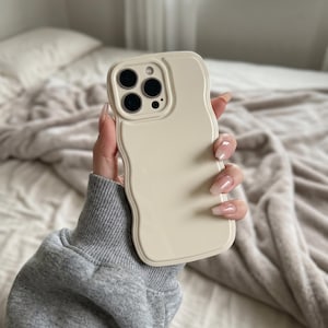 May include: A solid cream-colored phone case with a wavy, irregular edge design. The case features a raised rectangular camera cutout section with three circular lens openings and a small flash aperture, maintaining a smooth, matte finish.