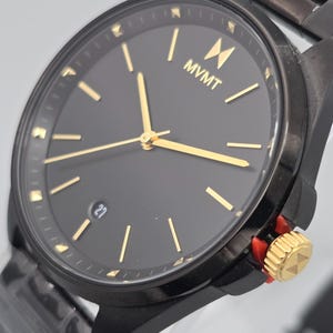 May include: A close-up shot of a black MVMT watch featuring gold hands and markers. The watch has a black strap and a gold-coloured crown with a red accent. The date is visible at the 6 o'clock position.