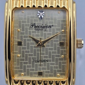 Puede incluir: Un reloj de pulsera rectangular dorado con una esfera texturizada y un acento en forma de diamante. La esfera del reloj muestra el texto "Precision Quartz" y "by GRUEN". El reloj tiene una correa dorada y una corona en el lateral.