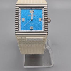 May include: A rectangular Invicta quartz watch with a light blue face and silver hands and numerals. The watch has a silver-colored, textured bezel and a matching textured band. The brand name "INVICTA" is visible on the watch face and band.