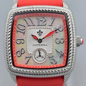 Op de afbeelding: Een zilverkleurig horloge met een vierkante wijzerplaat en een rode band. De wijzerplaat heeft een parelmoer achtergrond met zwarte cijfers en rode wijzers. De merknaam "JUDITH RIPKA" is zichtbaar op de wijzerplaat.