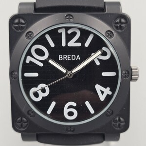 Puede incluir: Reloj de pulsera cuadrado negro con esfera negra y números blancos. La esfera del reloj muestra la marca "BREDA". El reloj tiene una correa negra y una corona plateada. La caja del reloj está asegurada con cuatro tornillos.