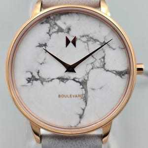 May include: A rose gold wristwatch with a white marble-effect face and black hands. The watch has a grey leather strap. The word "BOULEVARD" is printed on the face.