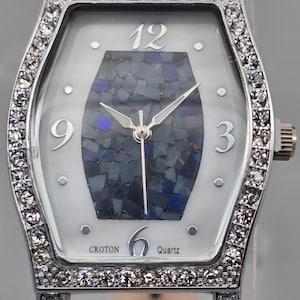 Puede incluir: Reloj de cuarzo plateado con esfera blanca y centro estilo mosaico. El reloj tiene un bisel rectangular incrustado con pedrería y una correa de cuero blanco. La esfera muestra los números 3, 6, 9 y 12.