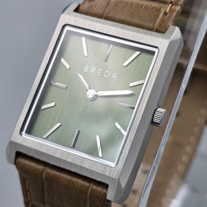 May include: A square-faced wristwatch with a silver case and a light green dial. The watch features the brand name "BREDA" and has a brown leather strap. The watch hands and markers are silver.