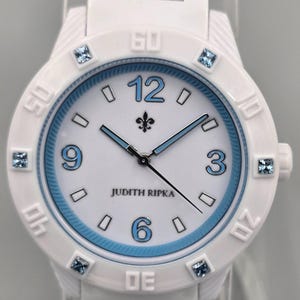 Puede incluir: Reloj de pulsera blanco con correa blanca y esfera blanca con detalles azules. La esfera del reloj muestra la marca "JUDITH RIPKA" y números azules a las 12, 3, 6 y 9. Cristales cuadrados azules marcan las horas.