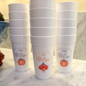 May include: A stack of white foam cups featuring a printed design with the text The Phillips. The design includes a shield-shaped border, a watercolor-style orange pumpkin, and a turkey wearing a hat on the center cup, all in orange and brown.