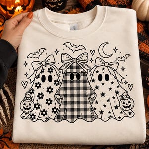 May include: A cream-coloured crewneck sweatshirt featuring a black line-art graphic of three ghosts wearing bows. The ghosts have floral, buffalo plaid, and star patterns. They are surrounded by bats, stars, hearts, and a crescent moon.