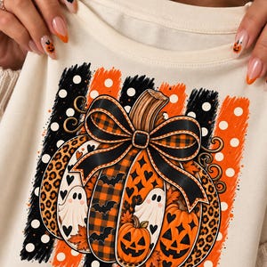 May include: A cream-coloured sweatshirt featuring a graphic print of a large pumpkin decorated with a black and orange plaid bow. The pumpkin design includes leopard print, ghost figures, small hearts, and jack-o'-lantern faces on a striped background.