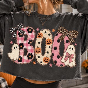Könnte beinhalten: Ein anthrazitfarbenes, langärmeliges bauchfreies Sweatshirt mit dem Wort BOO in großen Buchstaben. Das B ist rosa kariert mit einer schwarz-weißen Vichy-Schleife, das O ist cremefarben mit orangefarbenen Kürbissen und das O ist rosa gepunktet mit einer Leoparden-Schleife.