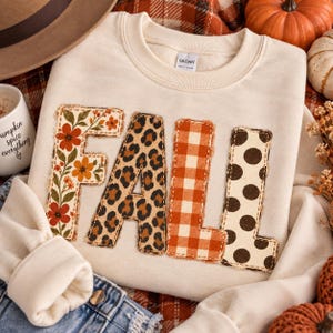May include: A cream-colored crewneck sweatshirt featuring the word FALL appliqued across the front. Each letter has a distinct pattern: the F is floral, the A is leopard print, the L is orange gingham, and the L is white with brown polka dots.
