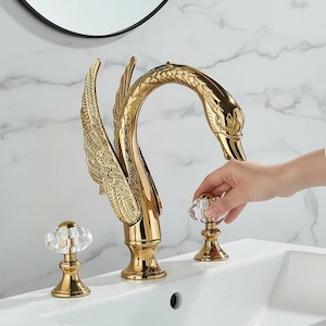 Può includere: Un rubinetto dorato a forma di cigno con pomelli di cristallo. Il rubinetto presenta dettagliati disegni di ali e testa di cigno. È installato su un lavandino bianco, con una parete a motivi marmorei sullo sfondo.