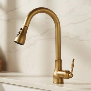 May include: A gold-toned kitchen faucet with a pull-down sprayer. The faucet has a curved spout and a lever handle. The design is modern and elegant, suitable for a contemporary kitchen. The faucet is set against a white countertop and a marble-patterned wall.