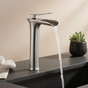 Puede incluir: Un grifo de baño de níquel cepillado con un caño en cascada. El agua fluye del caño a un lavabo negro. Una pequeña suculenta en una maceta negra se encuentra a la derecha del grifo. Un paño blanco está a la izquierda.