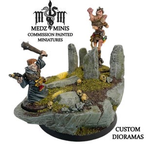 May include: A custom diorama with two miniature figures on a rocky base with skulls and stone pillars. One figure holds a club and a scope, the other is bound. The base is black, and the text "CUSTOM DIORAMAS" is visible.
