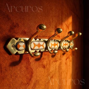 May include: A brass coat rack for wall mounting, with four curved hooks. The rack has decorative orange and white inlaid designs. The word "Arcuros" is visible in the top left and bottom right corners.