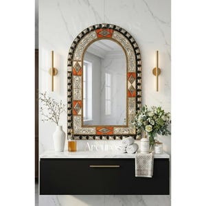 Op de afbeelding: Een boogvormige spiegel met een decoratief frame met oranje, zwarte en witte geometrische patronen. De spiegel is gemonteerd boven een zwarte wastafel met een wit aanrechtblad. De scène omvat een vaas, bloemen en andere badkameraccessoires.