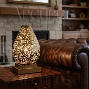 May include: A decorative table lamp with a teardrop-shaped shade made of perforated metal. The lamp sits on a square base and emits a warm glow. The lamp is on a wooden table.