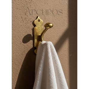 May include: A brass-coloured wall hook with an elaborate design, holding a white towel. The hook is mounted on a tan wall. The word "Arcuros" is printed in the upper left corner.
