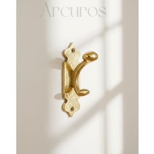 May include: A gold-colored wall hook with an ornate design. The hook has a decorative backplate with engraved patterns and two curved arms for hanging items. The word "Arcuros" is printed at the top.