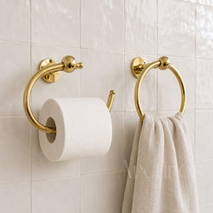 May include: A polished gold-tone metal toilet paper holder holding a roll of white toilet paper and a matching circular towel ring holding a beige linen towel, both mounted on a wall covered in square, glossy, off-white ceramic tiles.