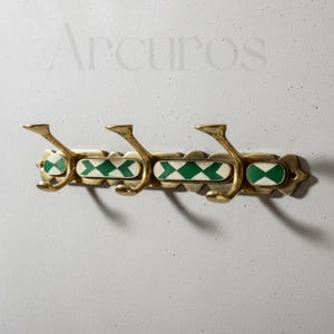 Puede incluir: Un perchero de pared de latón con tres ganchos. El perchero presenta un diseño decorativo con patrones de diamantes verdes y blancos. La palabra "Arcuros" es visible en el fondo.