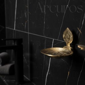 Pode incluir: Um saboneteira dourada com um design ornamentado, montada numa parede de mármore preto. A saboneteira tem uma placa traseira decorativa e uma base curva em forma oval. A palavra "Arcuros" está no topo da imagem.